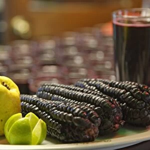 Peru, Lima, three purple maize corn cobs, a quince, and a sliced lime on a plate plate, glass of chicha morada in the background